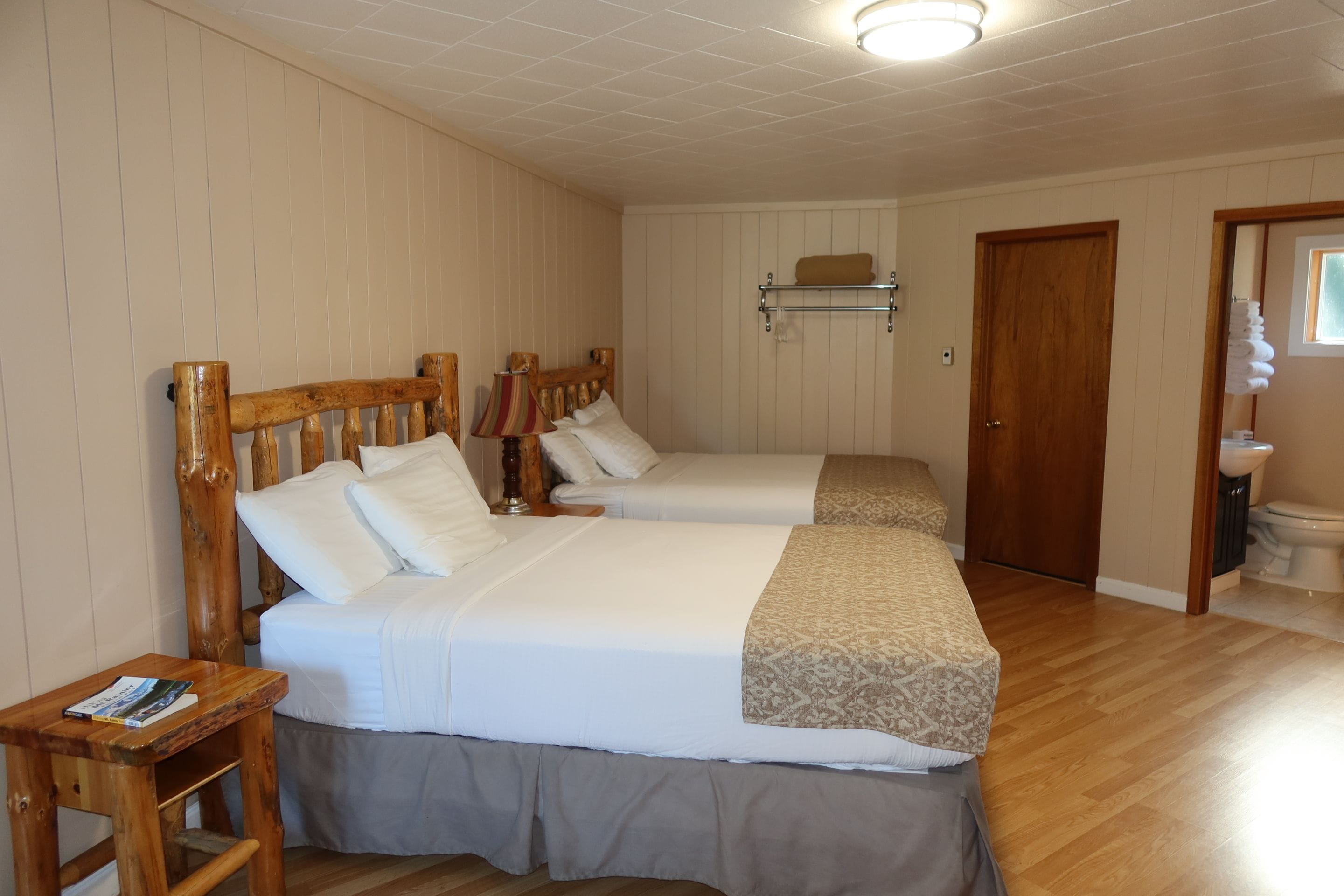 Habitación de hotel con dos camas, cabecero de madera, mesita de noche, lámpara, repisa con toallas y puerta de madera.