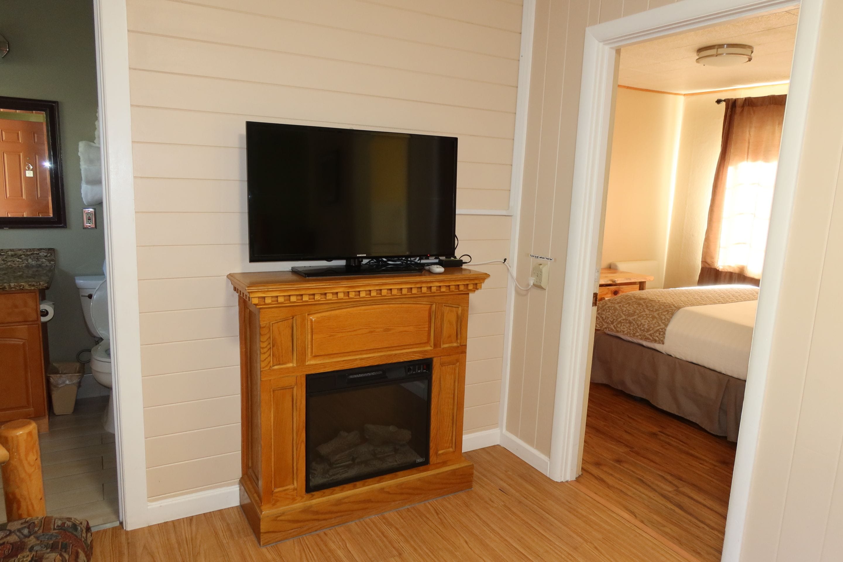A hotel room with a wooden fireplace, a television mounted above it, a bed, and a bathroom visible in the corner.