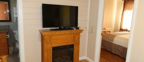 A hotel room with a wooden fireplace, a television mounted above it, a bed, and a bathroom visible in the corner.