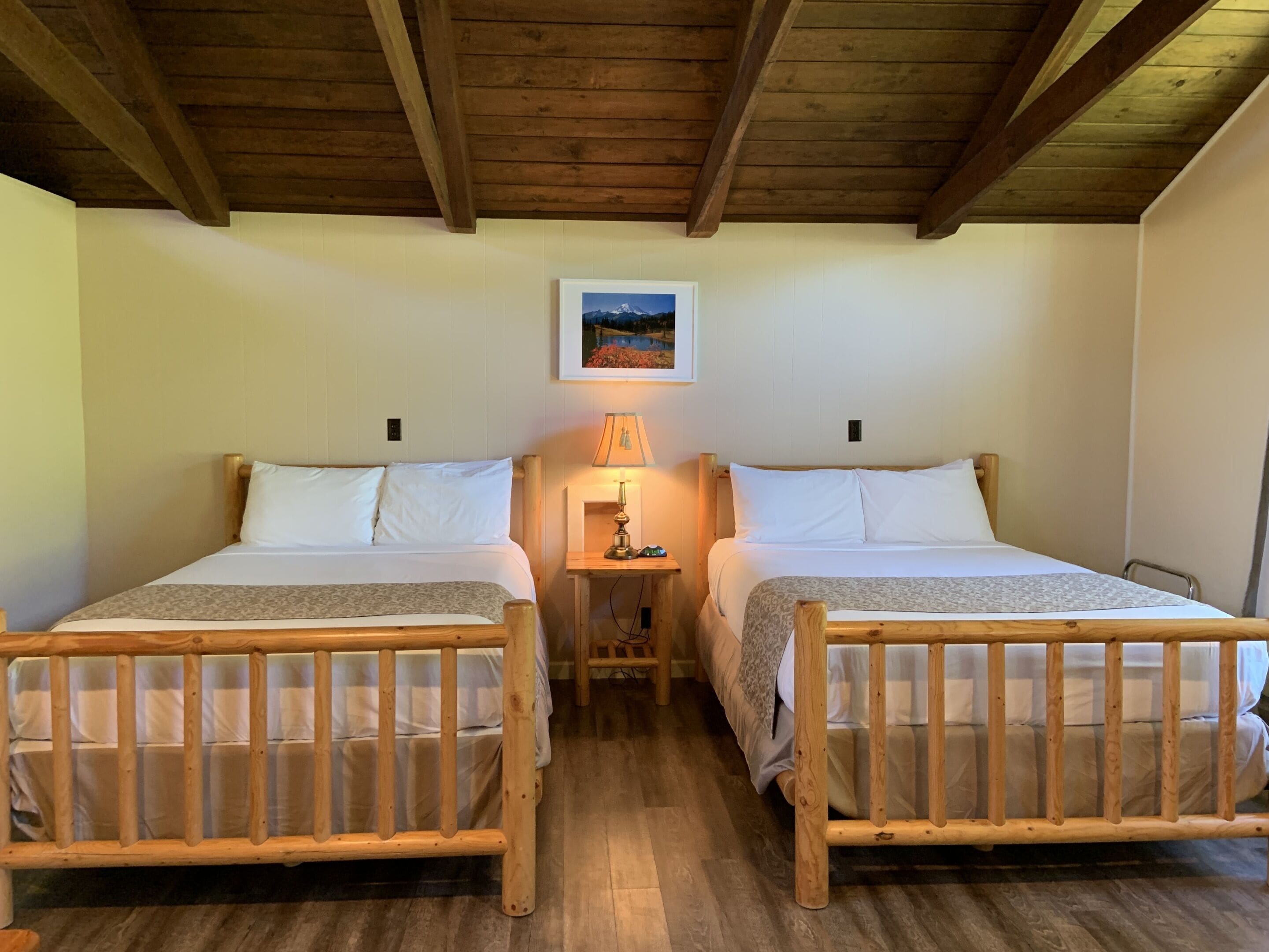 Een kamer met twee houten bedden, een nachtkastje met een lamp en een lijst met een afbeelding aan de muur.