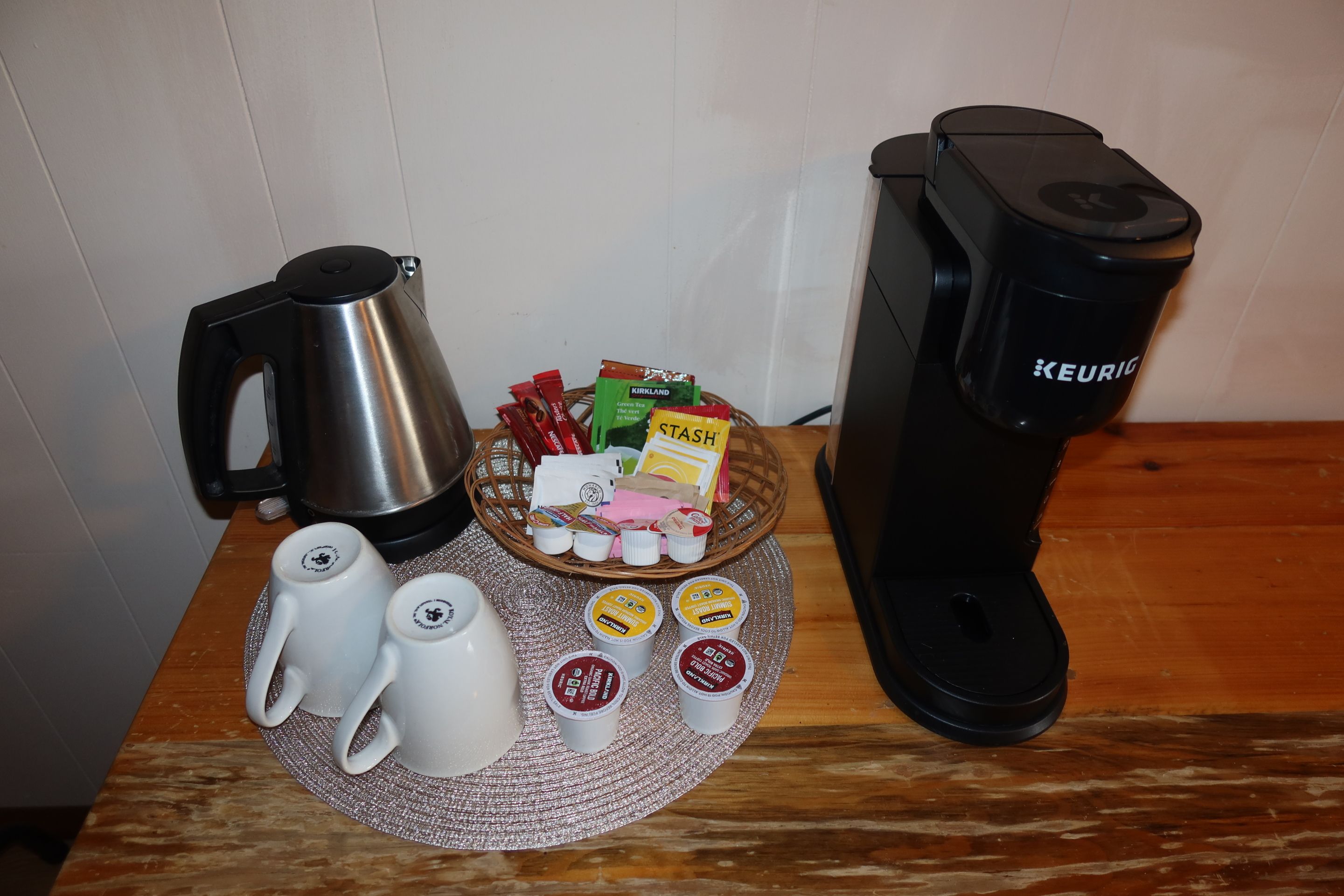 Una cafetera, un hervidor y una cesta con cápsulas de café y sobres de azúcar sobre una mesa de madera.