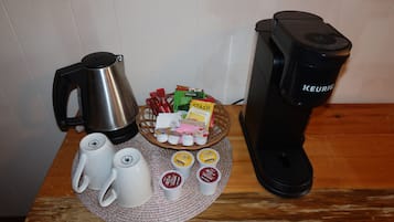 Une machine à café, une bouilloire et un panier contenant des dosettes de café et des sachets de sucre, le tout posé sur une table en bois.