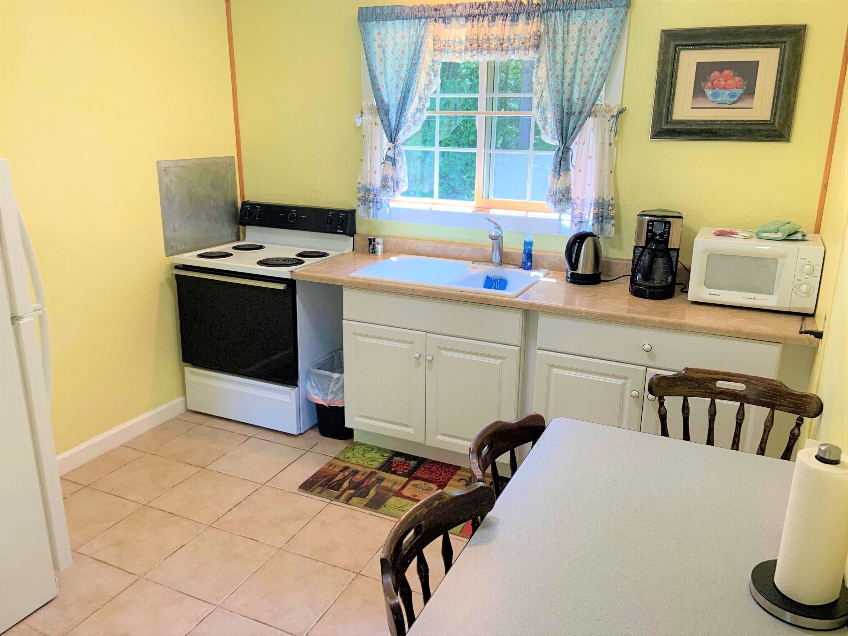 A small kitchen with a stove, oven, sink, and microwave. There is a window with curtains, a chair, and a table.