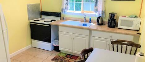 A small kitchen with a stove, oven, sink, and microwave. There is a window with curtains, a chair, and a table.