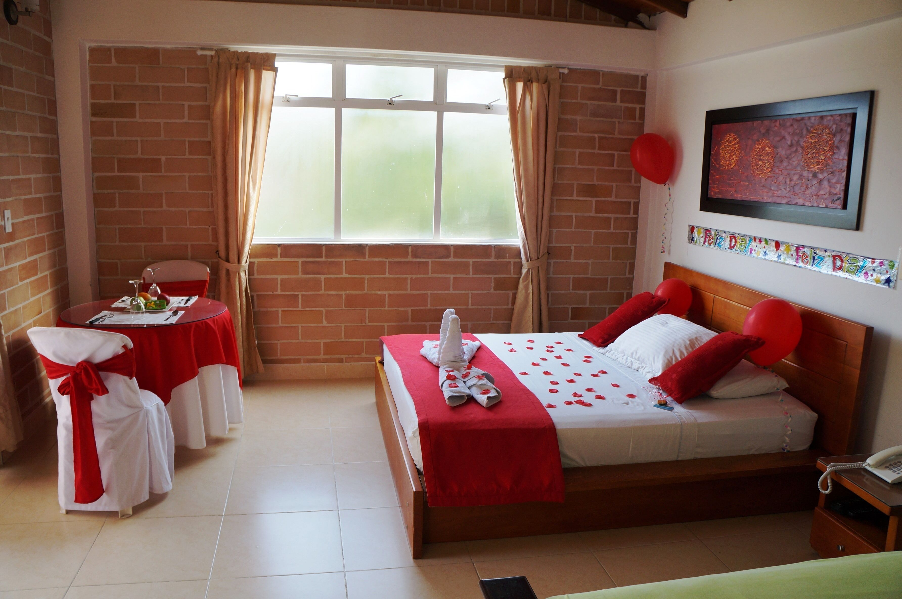 A hotel room with a brick wall, a bed with red and white bedding, a table with a red tablecloth, and a window with curtains.