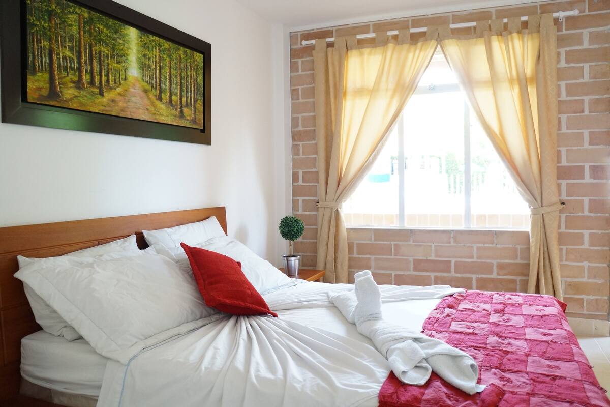 Un letto rifatto con lenzuola bianche, un cuscino rosso e una coperta rosa. Sopra il letto, un quadro raffigura un sentiero forestale.
