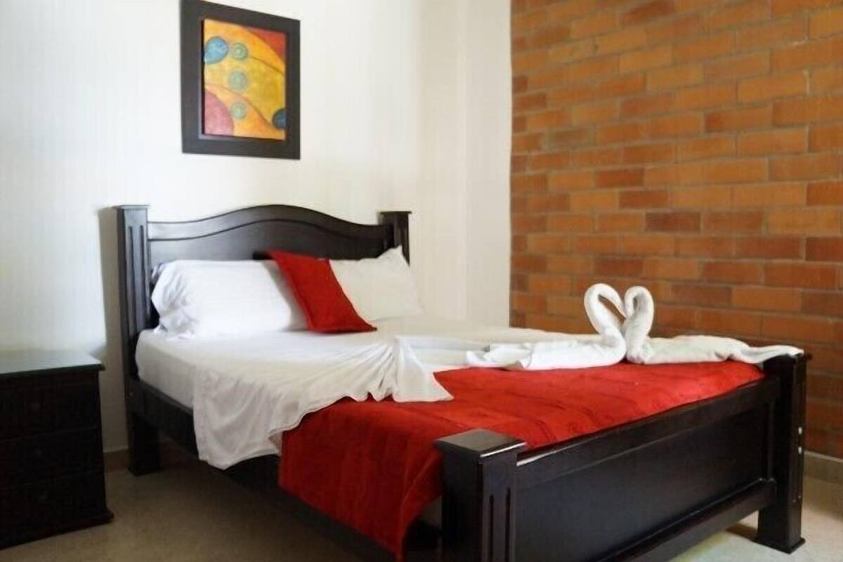 Un letto con coperta rossa, lenzuola bianche, poggiatesta nero e cuscino rosso.