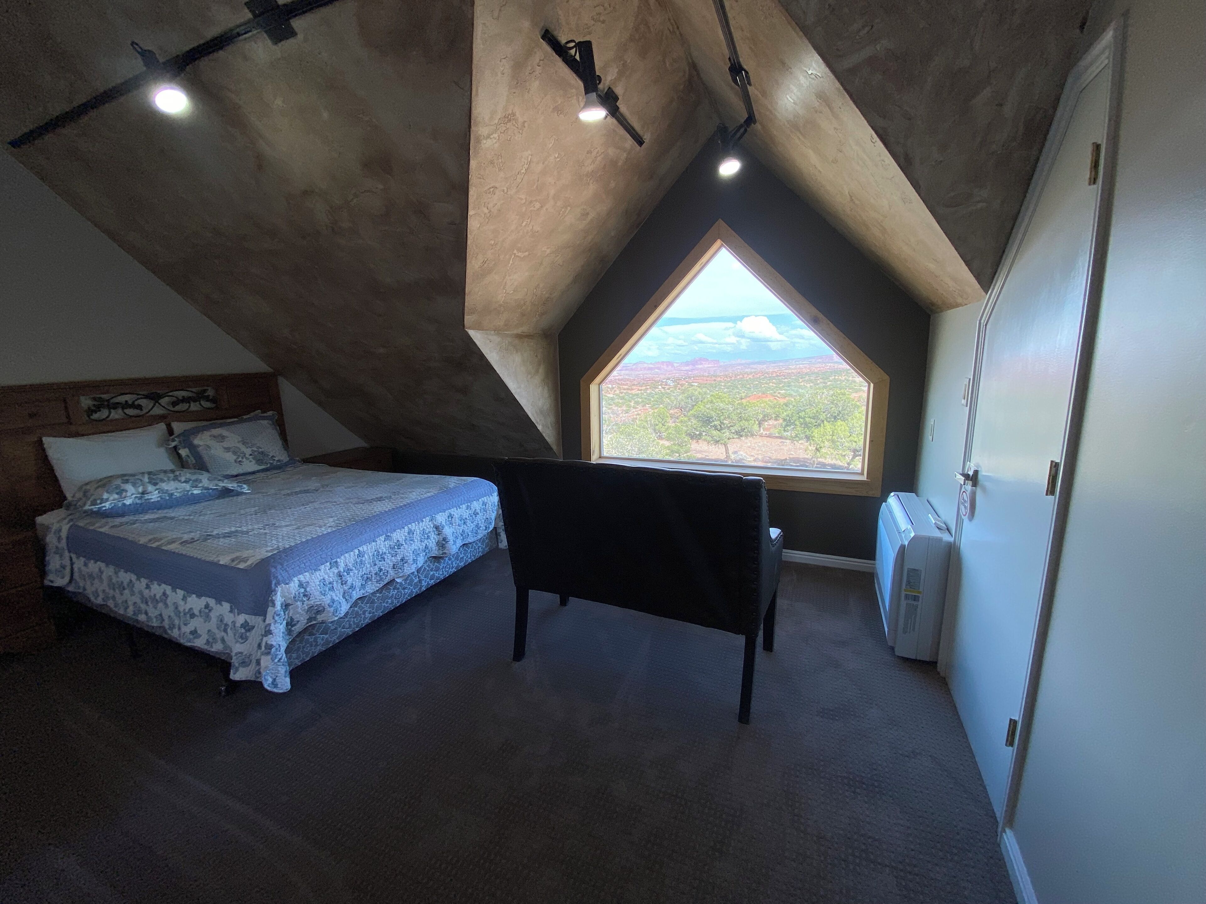 A bedroom with a sloped ceiling, a bed, a black chair, a radiator, and a window with a view of the landscape.