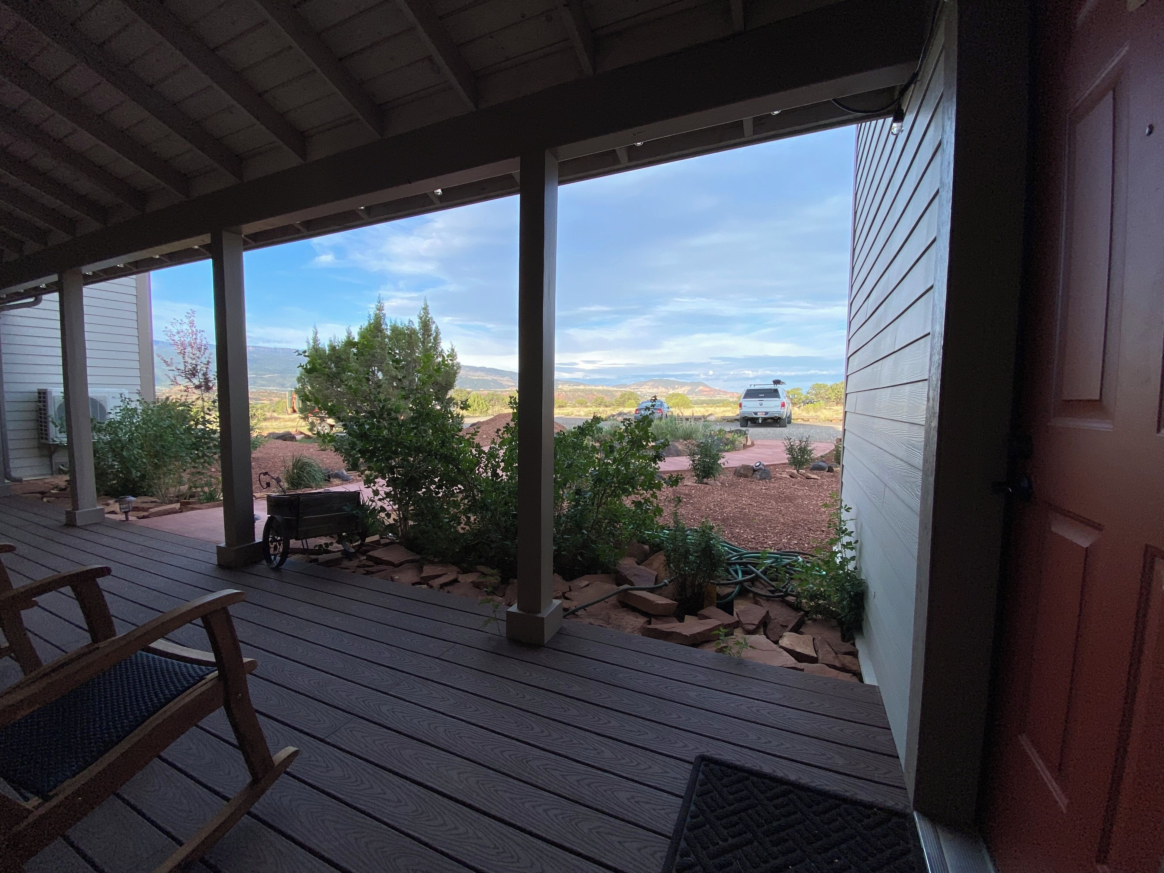 A covered porch with a view of a desert landscape, a white vehicle, and a wooden rocking chair.