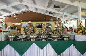 En bufféuppställning med tre serveringspersonal i uniform, ett buffébord täckt med en grön duk och en skylt med texten "Guaracu Hosteria" i bakgrunden.