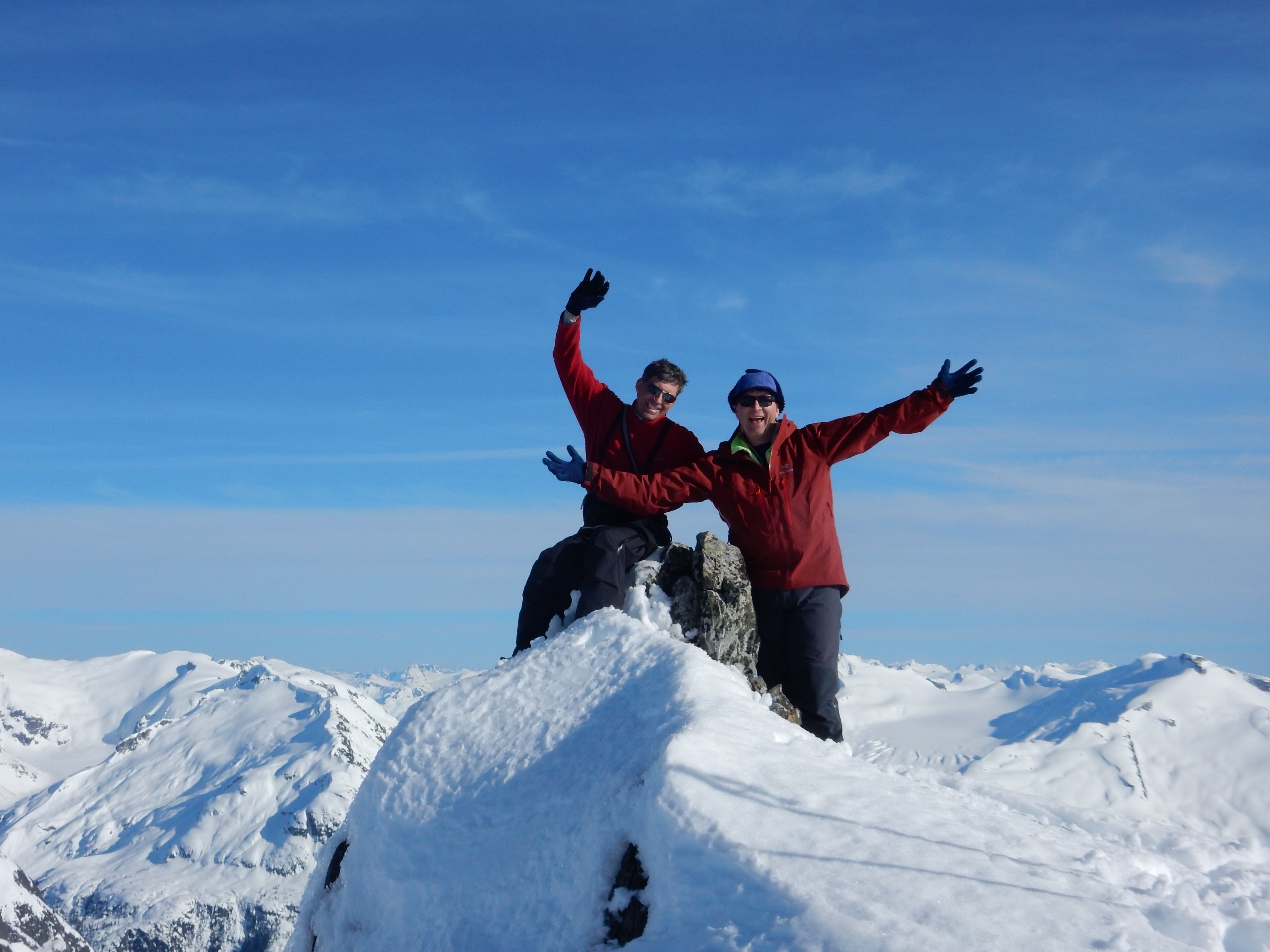 Two people standing on a snowy mountain peak with their arms raised, surrounded by snow-covered terrain and a clear blue sky.