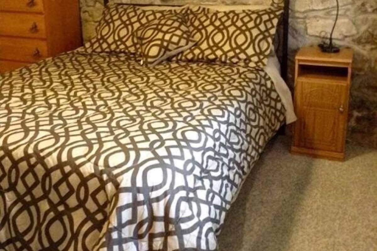 A bedroom with a stone wall, a patterned bedspread, a wooden dresser, and a nightstand with a lamp.