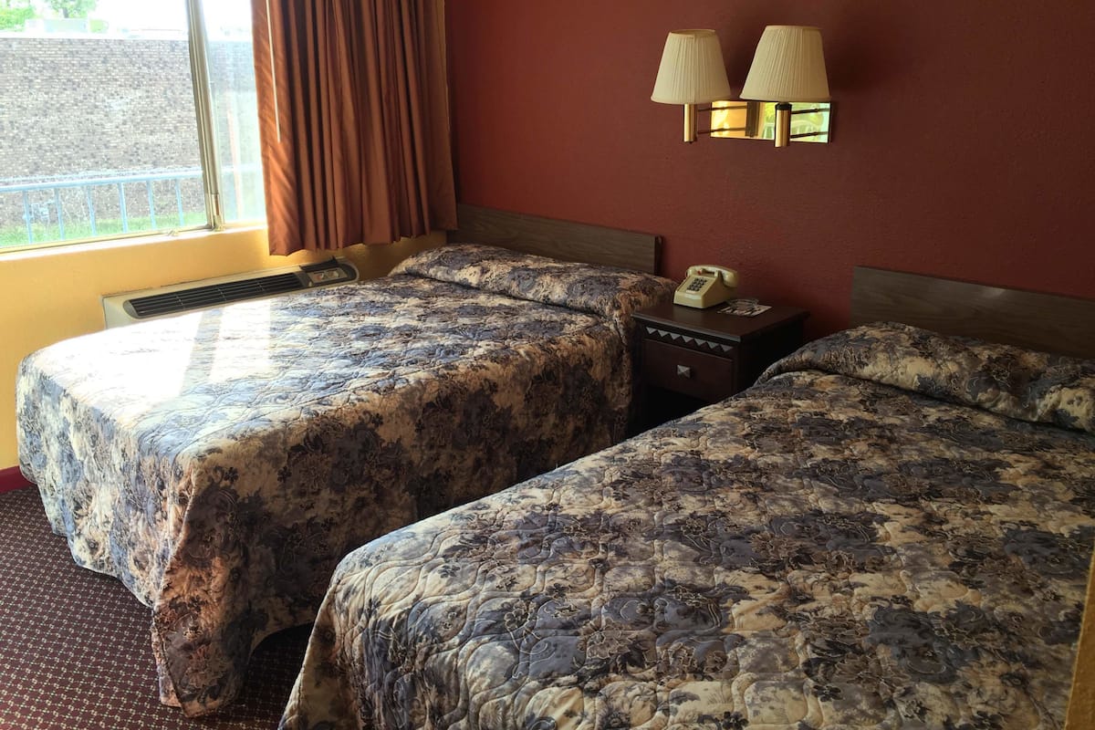 A hotel room with two beds, a window with curtains, a wall-mounted lamp, and a bedside telephone.