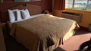 A hotel room with a large bed, a nightstand, a lamp, a window with curtains, and a view of the outside. - Super 7 Inn (Little Rock)