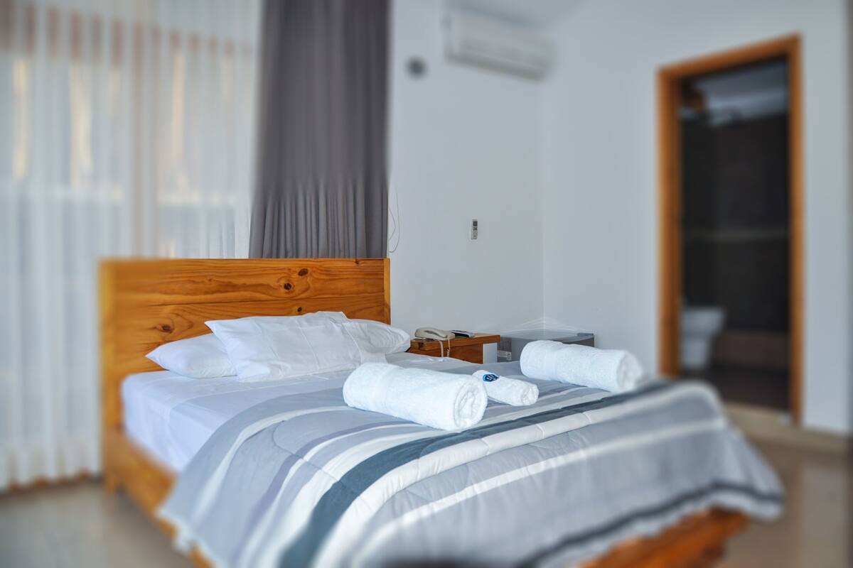 A neatly made bed with white linens and a striped blanket, a wooden headboard, a bedside table with a phone, and a window with curtains.