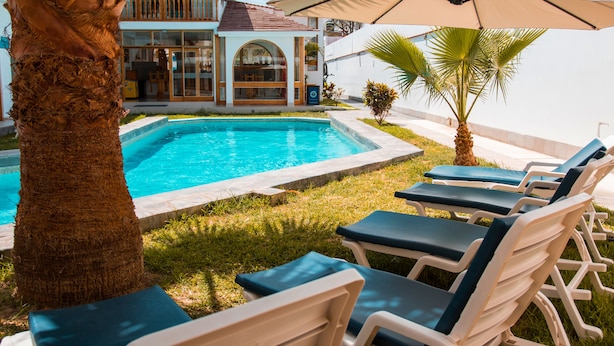 A pool area with lounge chairs and an umbrella, a palm tree, and a building with a balcony in the background.
