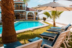 A pool area with lounge chairs and an umbrella, a palm tree, and a building with a balcony in the background. - Hotel Boutique La Angostura (Ica)