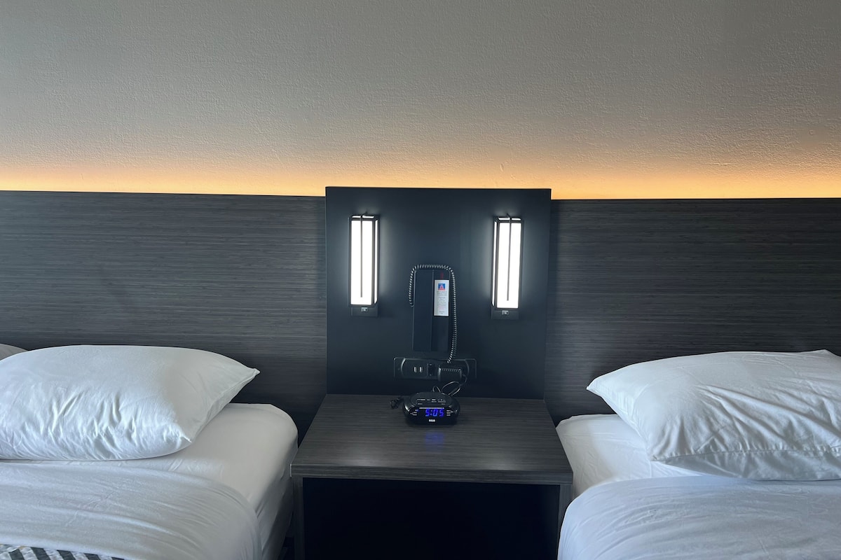 A hotel room with two beds, a nightstand, a wall-mounted phone, and a digital clock.