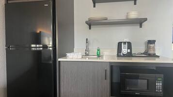 A compact kitchen with a black refrigerator, a microwave, a toaster, and shelves above the counter holding dishes and cups.