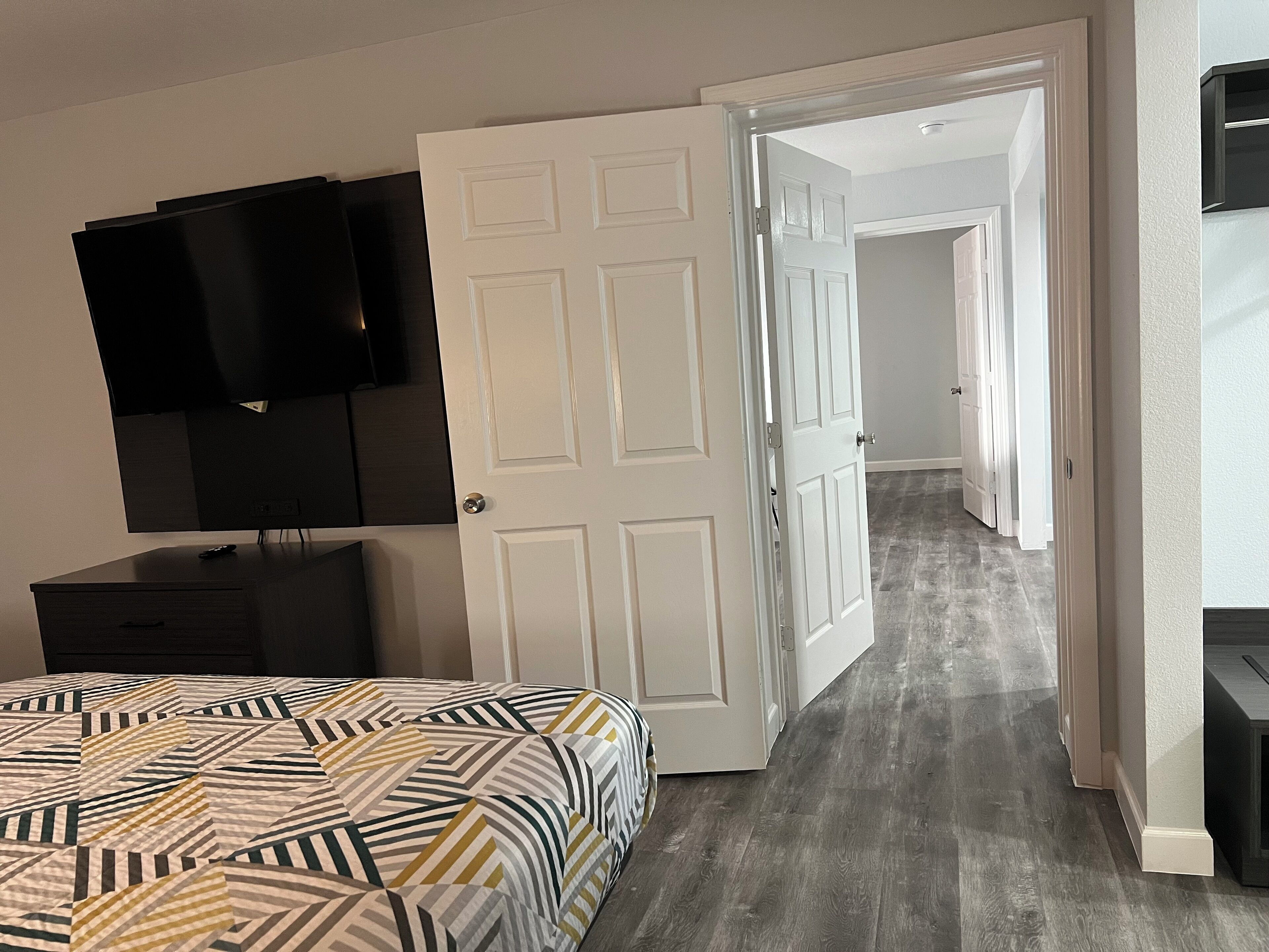 A bedroom with a bed, a TV mounted on the wall, and a door leading to a hallway.