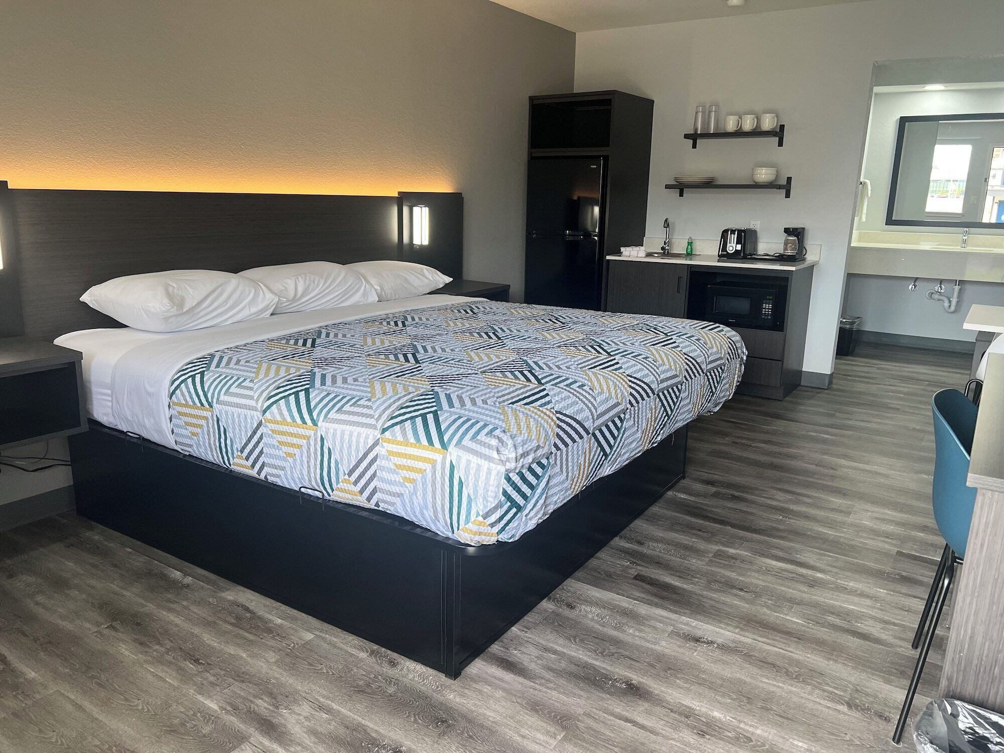 A modern hotel room with a large bed, a dark headboard, a bedside table, a small desk, a built-in coffee maker, and a mirror above the sink.