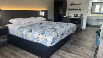 A modern hotel room with a large bed, a dark headboard, a bedside table, a small desk, a built-in coffee maker, and a mirror above the sink.