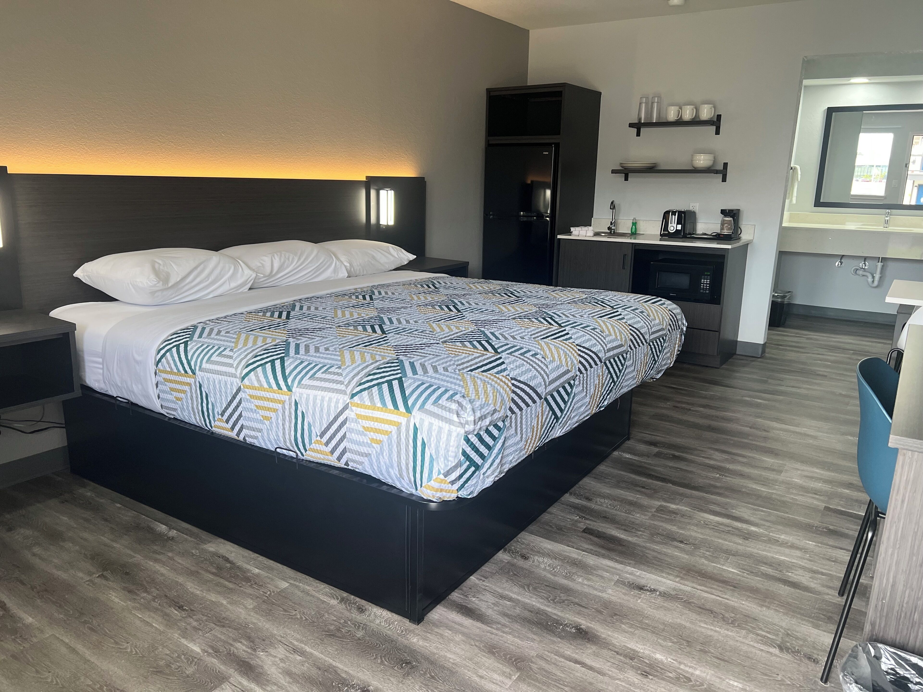 A modern hotel room with a large bed, a built-in desk, a kitchenette, and a bathroom.