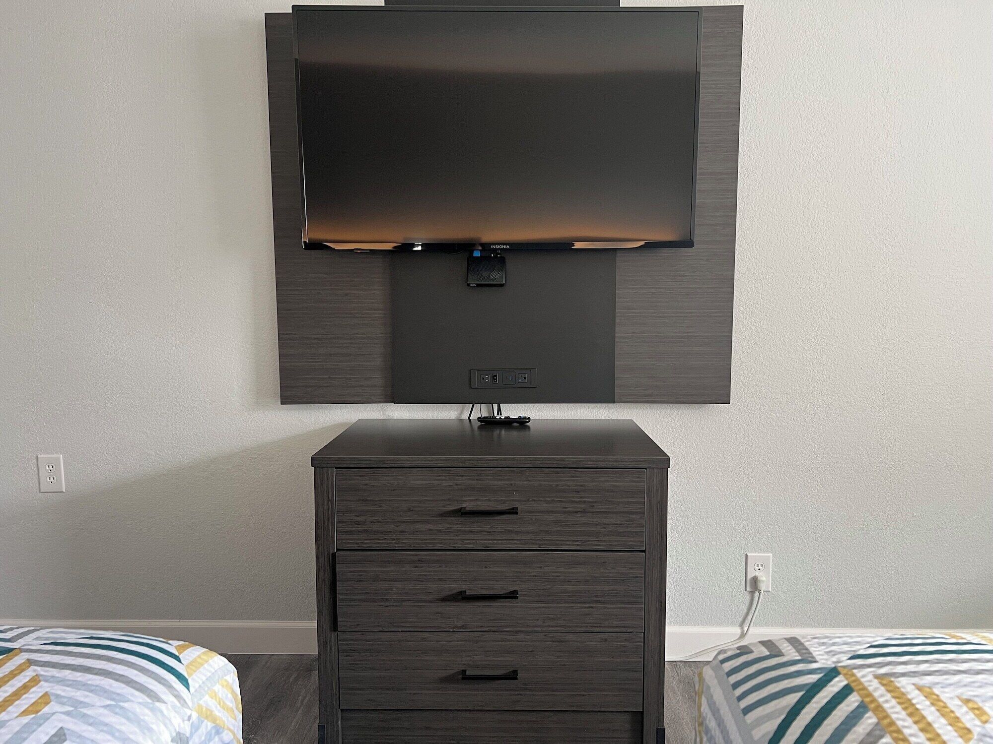 A room with a flat-screen TV mounted on the wall, a dark wooden dresser with drawers, and two beds with striped bedding.