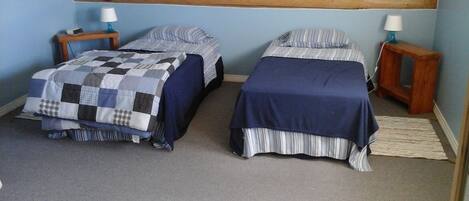 A bedroom with two single beds, each with a blue bedspread and matching pillows. There are bedside tables with lamps and a wooden headboard above the beds.