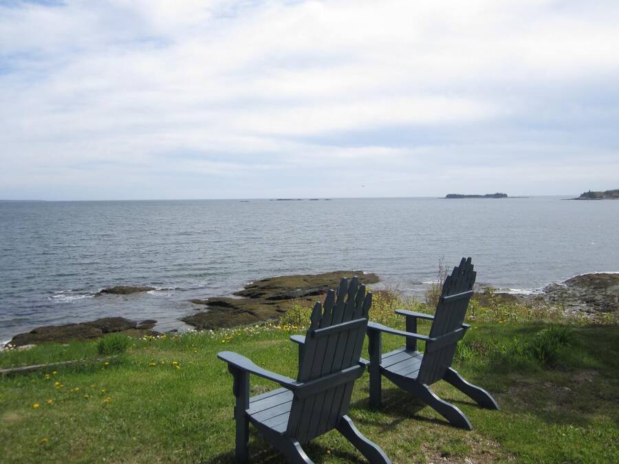 Two Adirondack chairs facing a rocky shoreline with a view of the sea and distant islands.