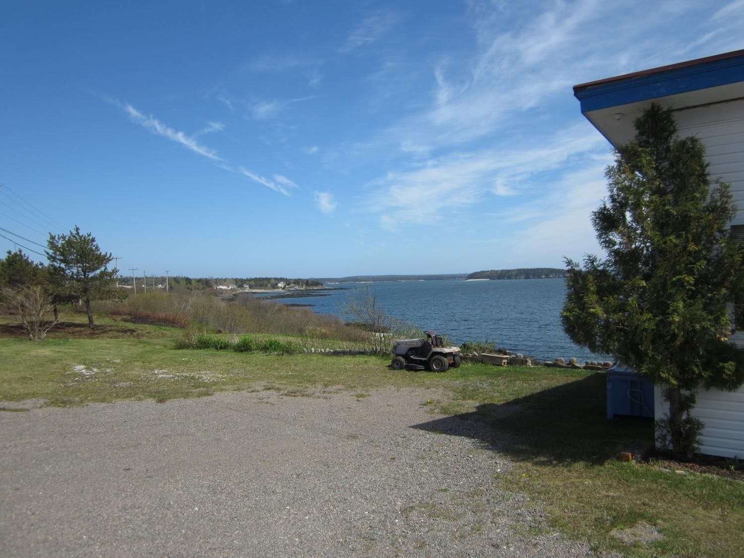 A gravel driveway leading to a building with a blue roof, overlooking a body of water and a distant shoreline.