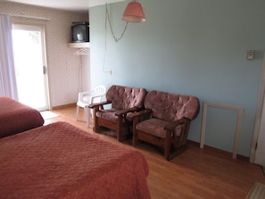 A room with two beds, a wooden sofa, a white chair, a television on a shelf, and a hanging lamp.