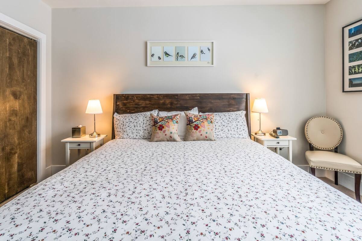 A bedroom with a large bed, two bedside tables with lamps, a chair, and a framed picture on the wall.