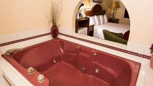Ein Hotelbad mit einer roten Badewanne, einem Spiegel, der ein Schlafzimmer reflektiert, und einer Vase mit Trockenpflanzen.