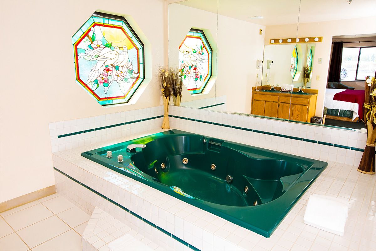 Un baño con una bañera verde grande, paredes cubiertas de azulejos blancos y dos ventanas de vidrio emplumado que representan pájaros.