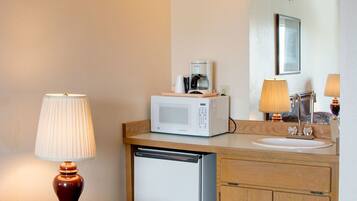 A hotel bathroom with a wooden vanity, a white sink, a microwave, a coffee maker, and a lamp.