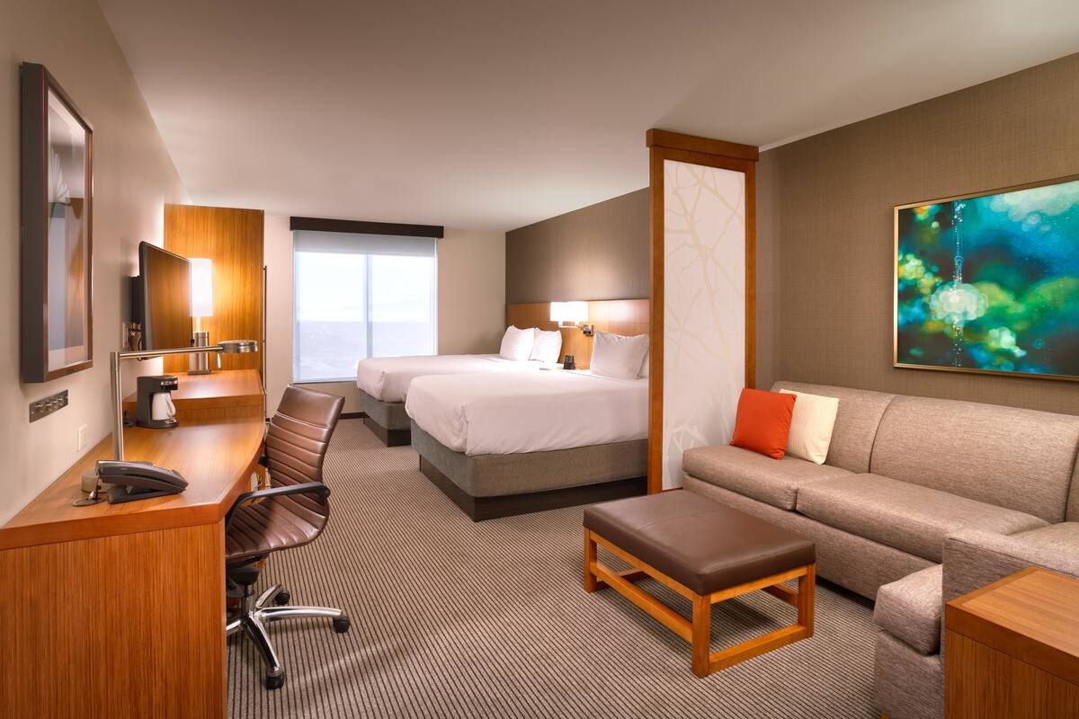 A hotel room with a desk, two beds, a sofa, and a large window.