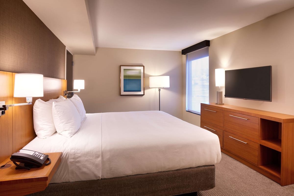A hotel room with a large bed, bedside lamps, a flat-screen TV, a wooden dresser, and a painting on the wall.