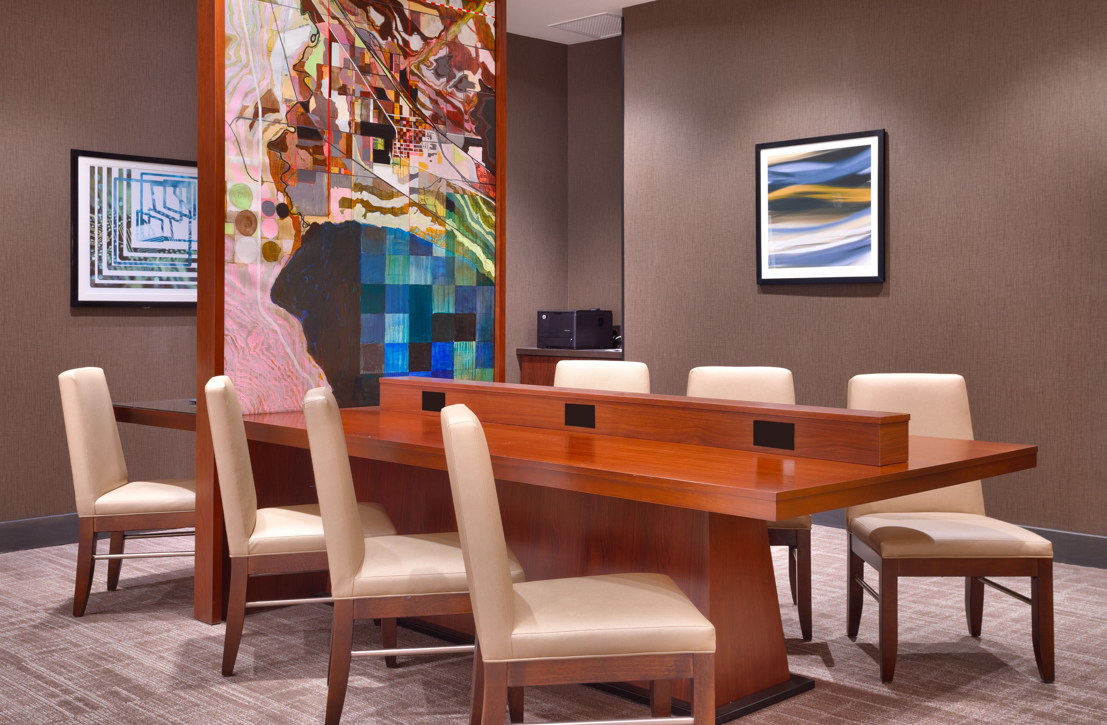 A conference room with a long wooden table, six beige chairs, and two framed abstract paintings on the walls.