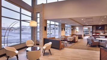 A spacious hotel lobby with large windows, a seating area with tables and chairs, and a reception desk.