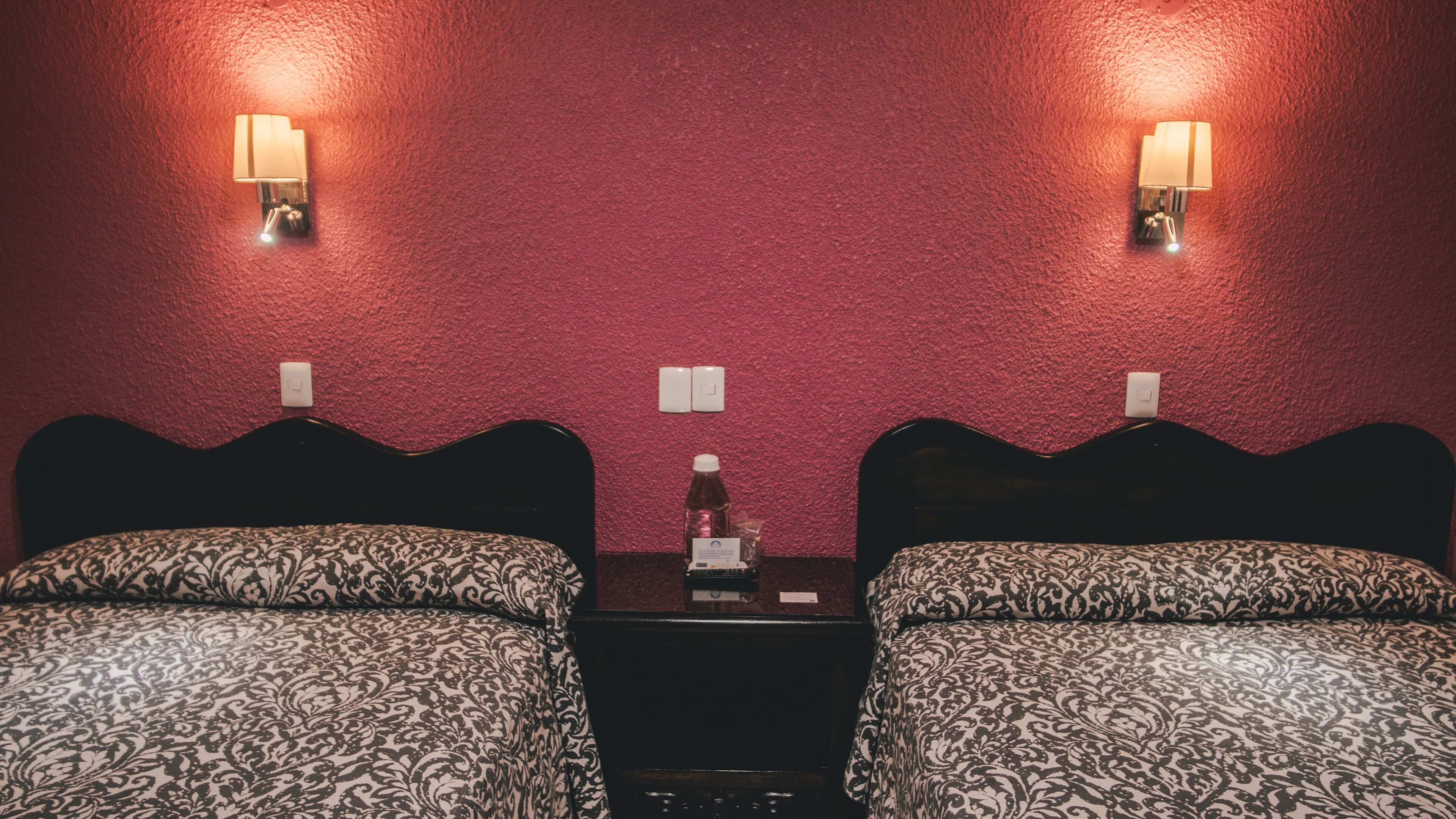 Habitación de hotel con dos camas, cada una con una colcha estampada, una mesita en el centro y lámparas fijadas a la pared.