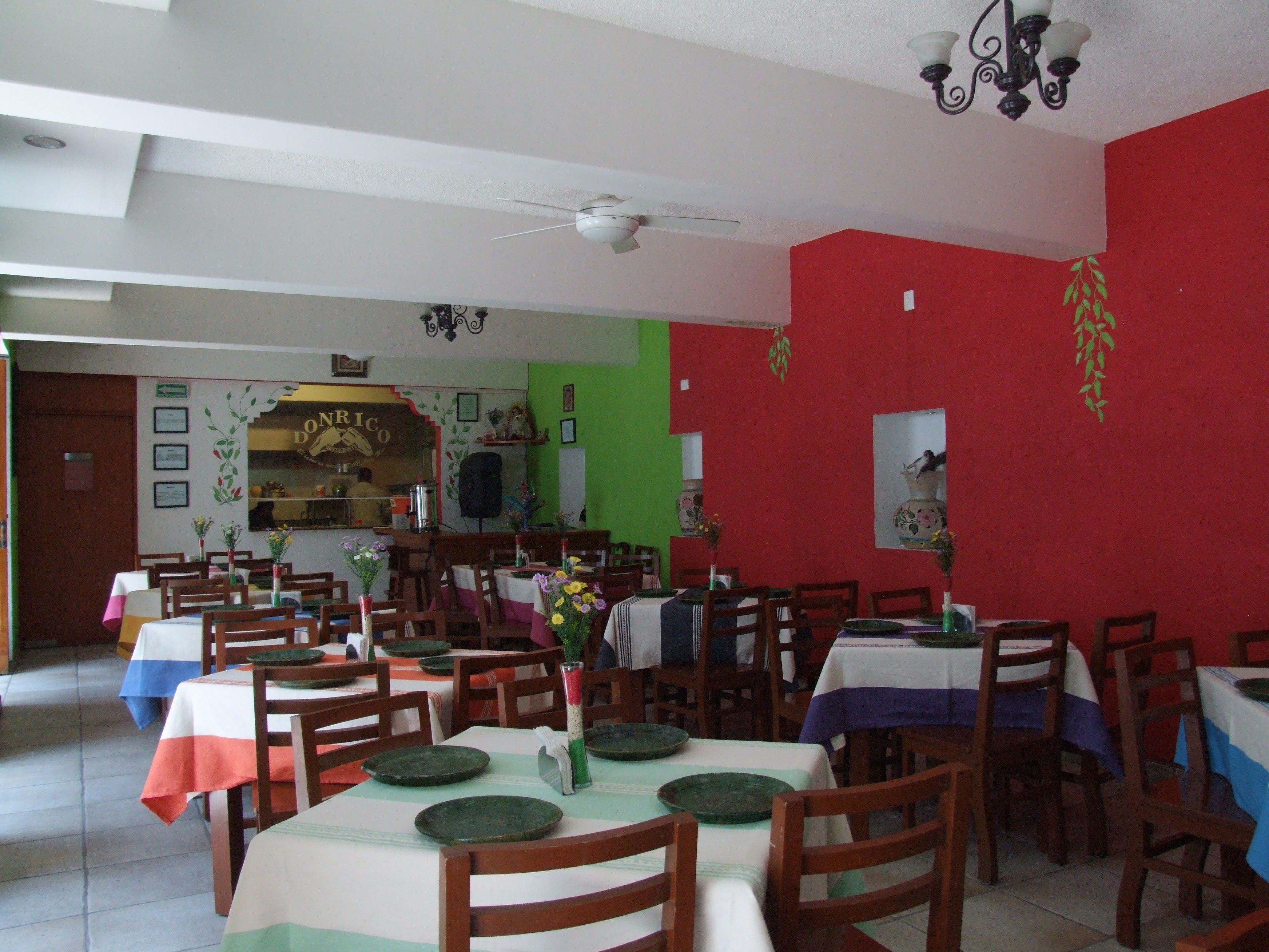 Un comedor con varias mesas y sillas, un ventilador de techo y una zona de cocina al fondo.