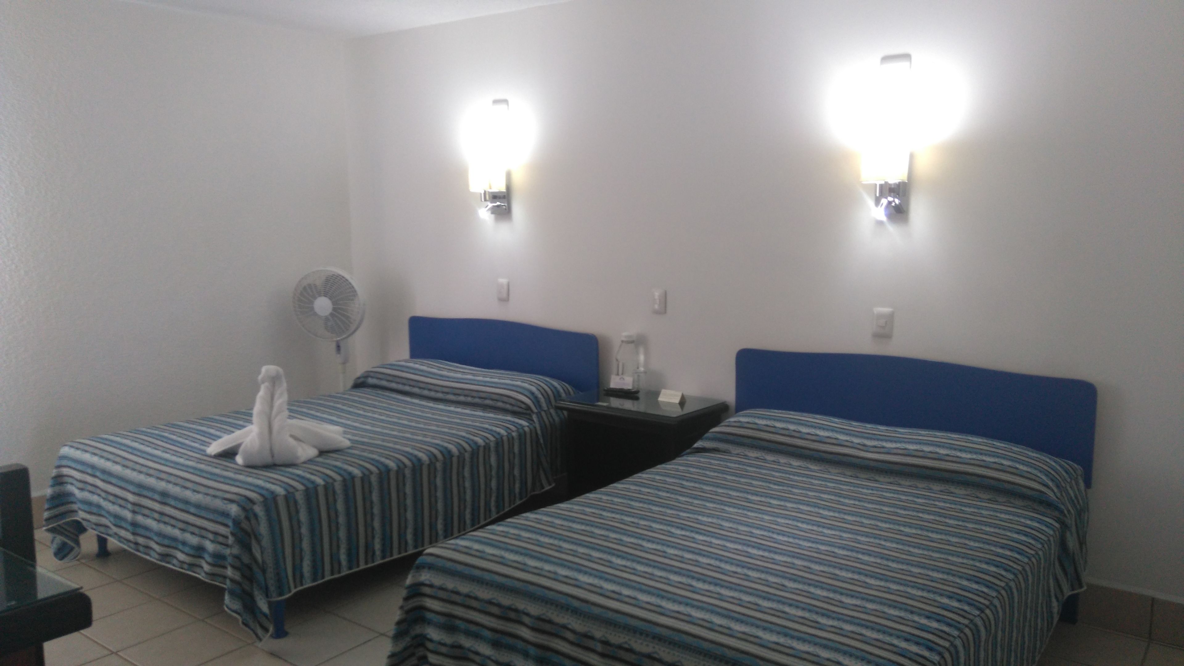 Une chambre d’hôtel avec deux lits simples, un ventilateur et deux lampes fixées au mur.