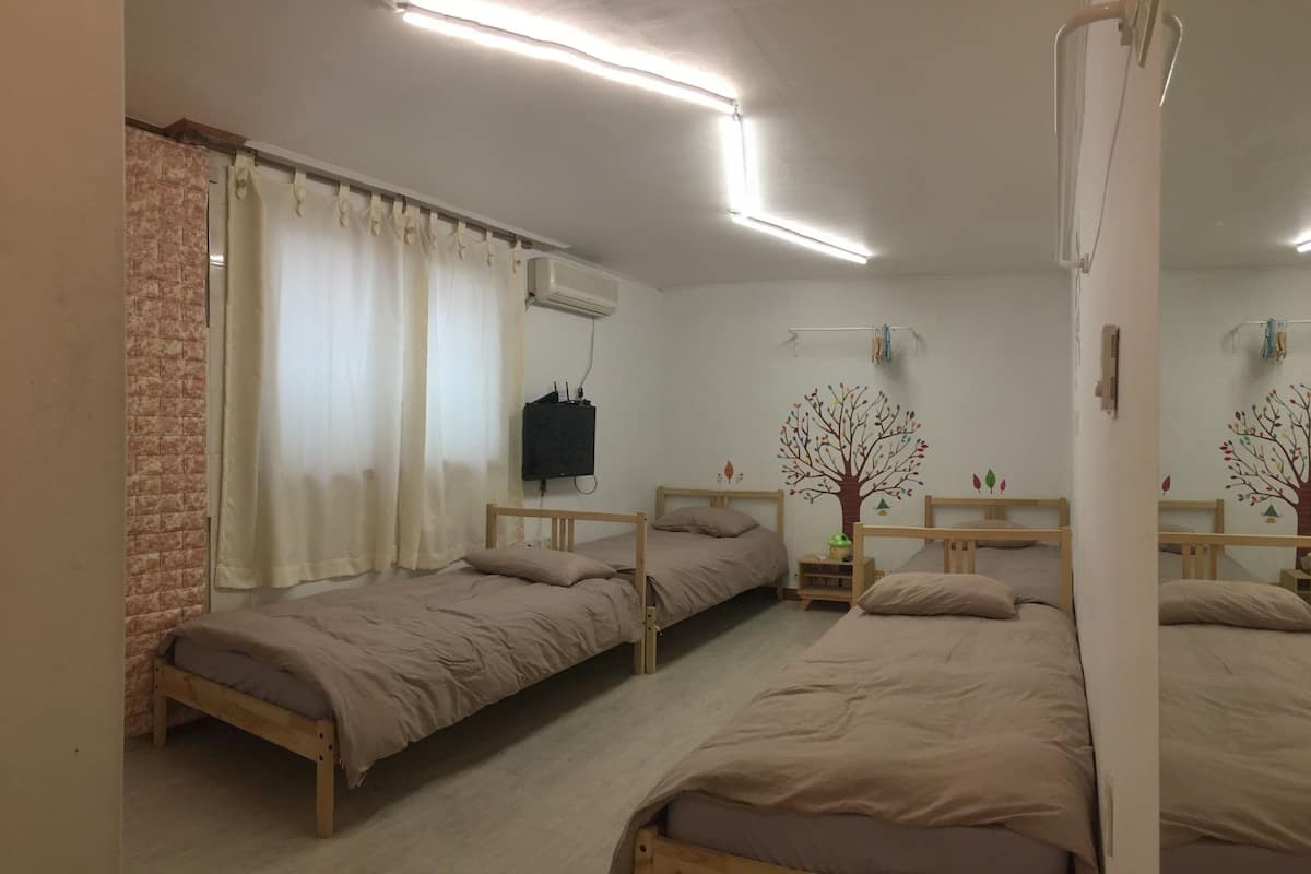 Une pièce avec trois lits en bois, un mur en briques, une fenêtre avec des rideaux, un appareil de climatisation et une étagère fixée au mur, sur laquelle se trouve une petite plante.