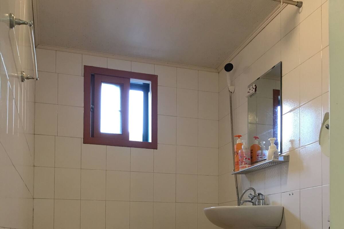 Une salle de bain avec des murs recouverts de carreaux blancs, une fenêtre, un lavabo et des toilettes.
