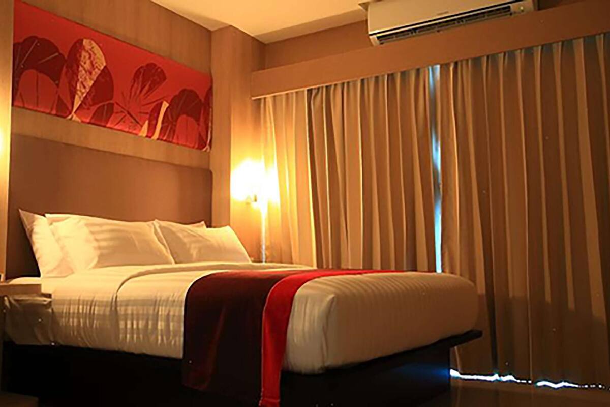 Una habitación de hotel con cama, dos lámparas de noche, aire acondicionado instalado en la pared y una ventana con cortinas.