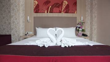 A neatly made bed with two white towels arranged in heart shapes, a red bedspread, and a floral painting above the bed.