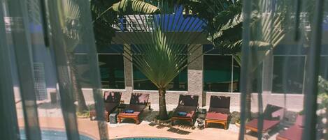 Zona de piscina con sillones reclinables y una palmera, rodeada por un edificio con amplias ventanas.