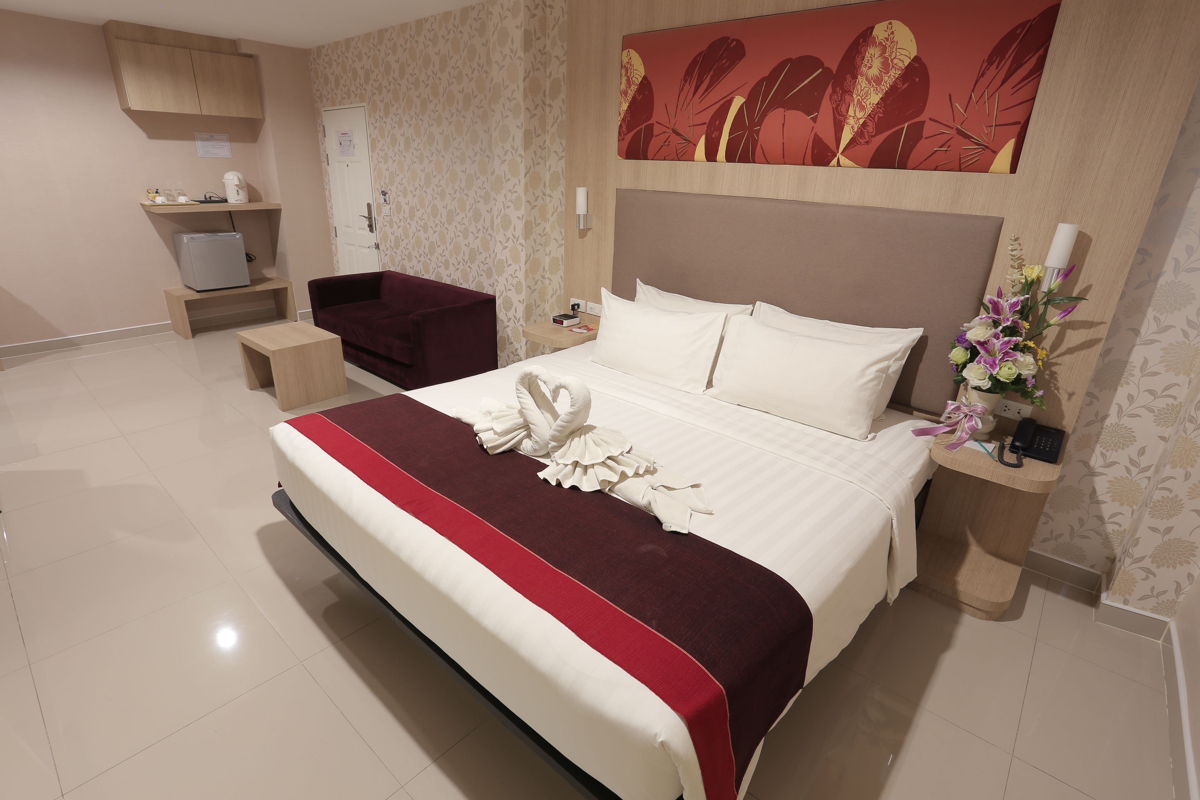 A hotel room with a large bed, a desk, a chair, a small refrigerator, a nightstand, and a wall-mounted artwork.