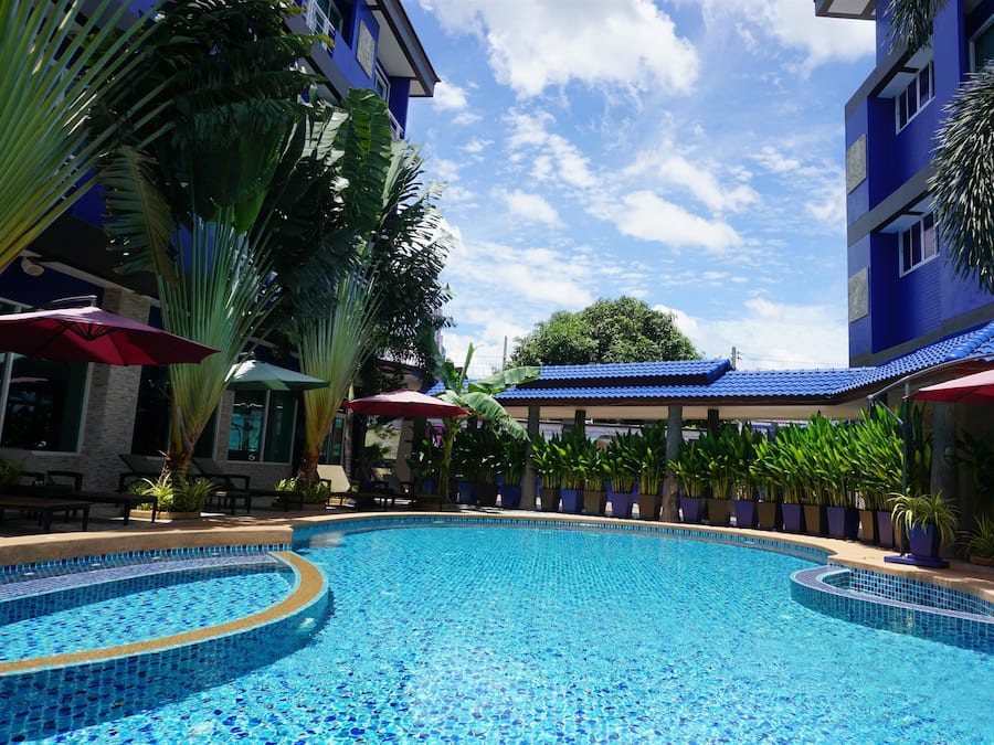 A swimming pool with tiled edges, surrounded by potted plants and umbrellas, with a building in the background.